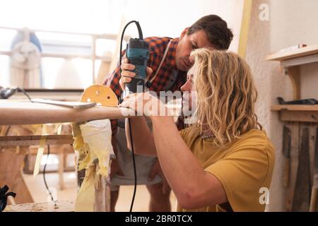 Zwei kaukasische männliche Surfboard Hersteller machen ein hölzernes Surfbrett zusammen mit einem Sander Stockfoto