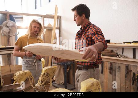 Zwei kaukasische männliche Surfboard Hersteller machen ein hölzernes Surfbrett zusammen und inspizieren es Stockfoto