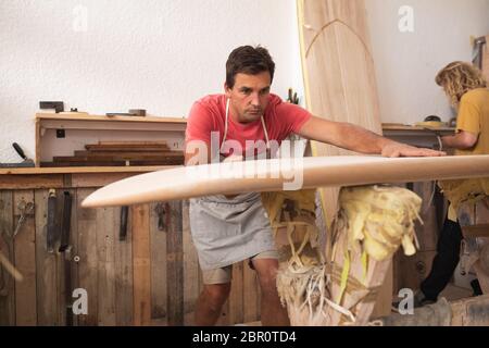 Zwei kaukasische männliche Surfboard Hersteller machen ein hölzernes Surfbrett zusammen und inspizieren es Stockfoto