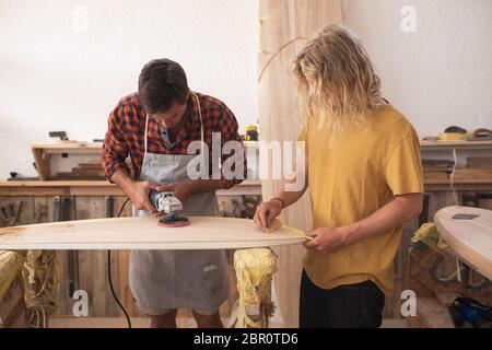 Zwei kaukasische männliche Surfboard Hersteller machen ein hölzernes Surfbrett zusammen mit einem Sander Stockfoto