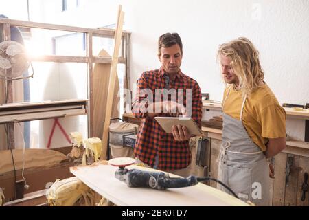 Zwei kaukasische männliche Surfboard-Hersteller diskutieren das Projekt und mit einem Tablet-Computer Stockfoto