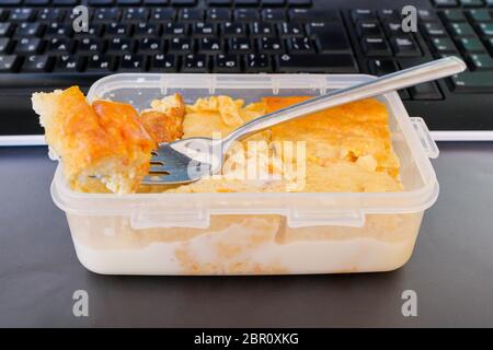 Appetitliche Hüttenkäse Kasserolle mit Kürbis in einem Kunststoffbehälter und einer Metallgabel auf einem schwarzen Computer-Tastatur Hintergrund. Mittagessen im Büro. Stockfoto