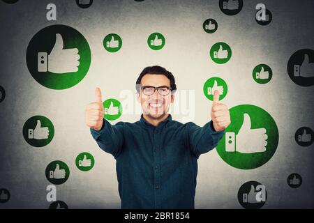 Fröhlicher Geschäftsmann zeigt Daumen nach oben positive Geste, schaut auf Kamera isoliert über graue Wand. Business Person trägt Brillen, lächelnd breit, a Stockfoto
