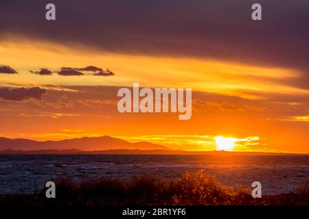 Die Sonne geht in den Westen, teilweise versteckt hinter lockigen Wolken, in der Nähe des Fischerdorfes Porto Lagos, Nordgriechenland, unter. Bergkette und Meerwasser in der Dunkelheit. Verwischte Büsche im Vordergrund Stockfoto