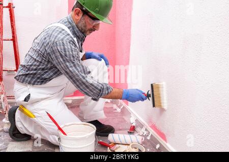 Kaukasische Haus Maler Arbeiter in weißen Overalls, mit Helm und Schutzbrille Malerei die rosa Wand mit weißen mit dem Pinsel malen. Bau indust Stockfoto