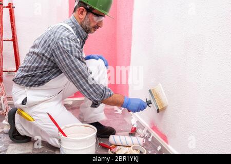 Kaukasische Haus Maler Arbeiter in weißen Overalls, mit Helm und Schutzbrille Malerei die rosa Wand mit weißen mit dem Pinsel malen. Bau indust Stockfoto