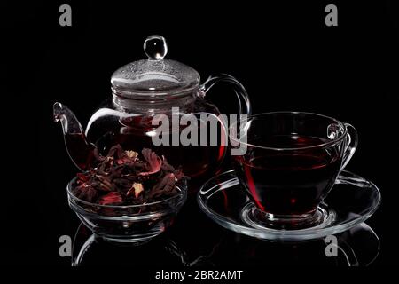 Hibiskus Tee in einem Glas Teekanne und Tasse, getrocknete Blütenblätter in Glasschale auf schwarzem Hintergrund. Stockfoto