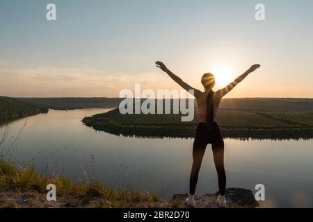 Silhouette einer sportlichen jungen Frau, die auf einem Bergrücken steht und den Sonnenuntergang über einem Flusstal genießt Stockfoto