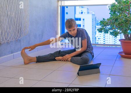 Kind mit Tablet-Computer, der Sportübungen macht, Yoga auf dem Balkon praktiziert. Sport, gesunder Lebensstil, aktive Freizeit zu Hause Stockfoto