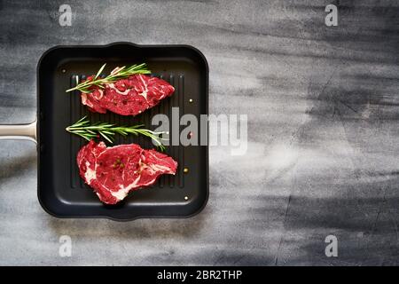 Zwei frische rohe Rindersteaks mit Rosmarin und Pfeffer in der Pfanne auf grauem Betongrund. Draufsicht, kopieren Sie Platz für Text. Stockfoto