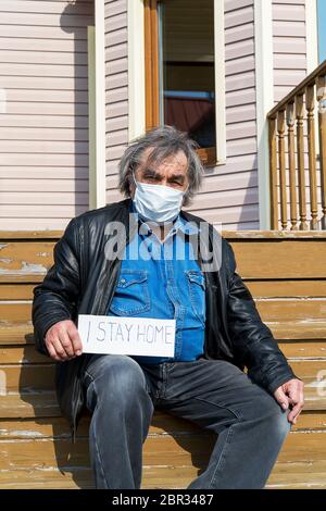 Virenschutz. Coronavirus Covid-19. Ein älterer älterer Mann in einer schützenden medizinischen Maske sitzt im Hof seines Hauses und schaut auf das kam Stockfoto