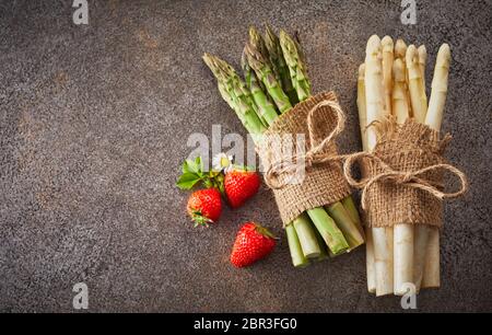 Zwei Bündel von frischen weißen und grünen Spargelspitzen liegen auf einem strukturierten grauer Hintergrund mit Kopie Raum und drei reife Erdbeeren Stockfoto