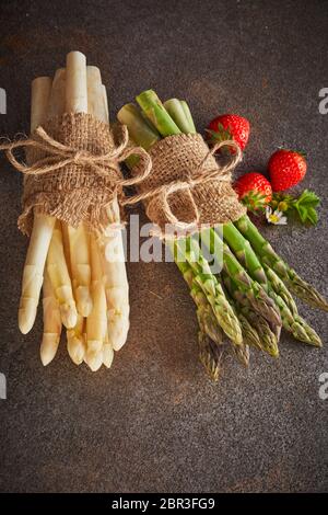 Frische rohe grüne und weiße Spargelspitzen in Bundles mit sackleinen auf einem grauen strukturierten Hintergrund mit drei reife rote Erdbeeren in vertikaler Form gebunden Stockfoto