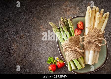 Trauben von frischem weißem und grünem Spargel mit hessischen und String auf einem Teller mit Erdbeeren über einen grauen Stein Hintergrund mit Copyspace gebunden Stockfoto