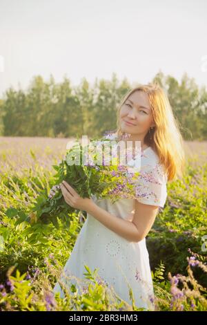 Junge Frau in weißem Kleid auf einer Wiese. Sie steht mit Blumenstrauß von wild lila Blumen in den Händen. Stockfoto