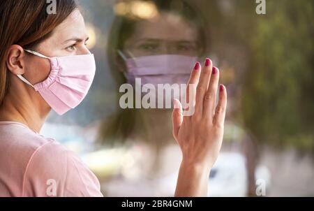 Junge Frau in Virusmaske sieht traurig aus dem Fenster hinter Glasscheibe, berühren sie mit der Hand. Quarantäne oder zu Hause bleiben, um sicher zu sein während der coronavir Stockfoto