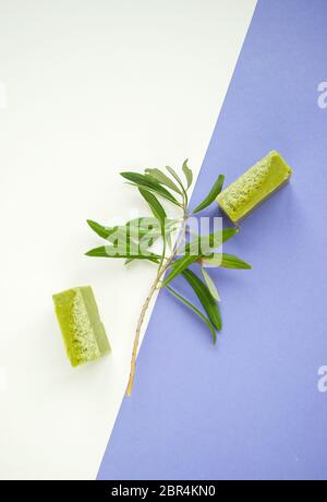Flaches Lay eines Olivenzweiges, handgemachte Olivenseifenstäbe auf purpurem und weißem Hintergrund. Stockfoto