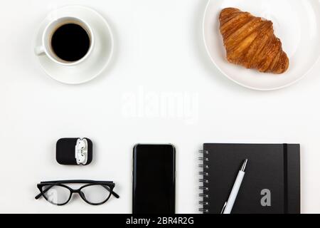 Moderne Arbeitsräume von Freiberufler. Smartphone, Kaffee, Notebook, Stift, Croissant, Gläser, Kopfhörer in Hülle auf weißem Hintergrund, Kopierraum, flach l Stockfoto