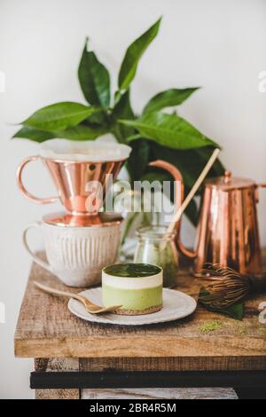 Kaffee und Dessert. Grüner Matcha-Käsekuchen und gebrauter schwarzer Kaffee in Kupferkanne auf rustikaler Holzküchentheke, weißer Wandhintergrund, schließen Stockfoto