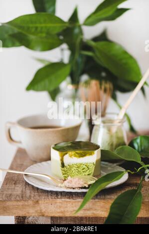 Kaffee und Dessert. Grüner Matcha-Käsekuchen und schwarzer Kaffee im Becher auf rustikaler Holzküchentheke, weiße Wand und Pflanze im Hintergrund, nah- Stockfoto