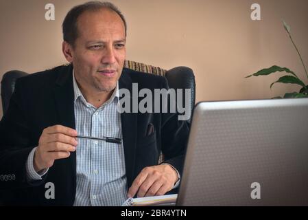 Intelligenter älterer Mann sitzt auf dem Sessel und arbeitet von zu Hause aus mit seinem Laptop, chattet mit seinen Kollegen und macht Notizen, mit einer Pflanze auf dem Bac Stockfoto