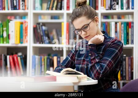 Mädchen sitzt in der Bibliothek. Junge Studentin in Brille im karierten Hemd sitzt in einer öffentlichen Bibliothek und liest ein Buch. Frau liebt es, Lesen zu entwickeln Stockfoto