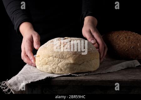 Weibliche Hände halten Runde gebackenen Weizen Mehl Brot über Holztisch, schwarzer Hintergrund Stockfoto