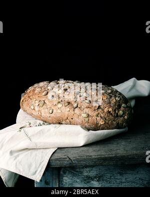 Gebackene oval Brot aus Roggen Mehl mit Kürbiskernen auf grauem Leinen Servietten, schwarzes Papier Hintergrund Stockfoto