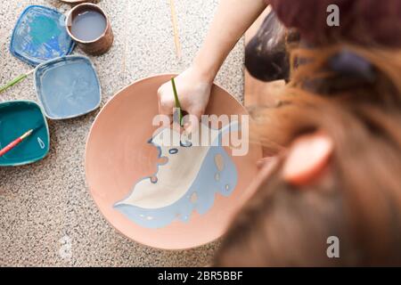 Potter Frau malt eine Keramikplatte. Mädchen zeichnet mit einem Pinsel auf Steingut. Prozess der Herstellung von Tonprodukten. Stockfoto