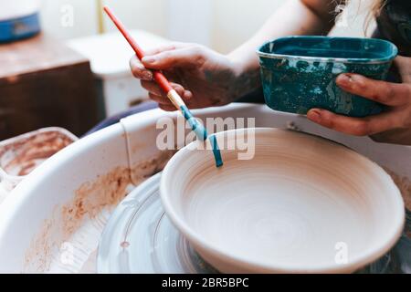 Professionelle Töpfer arbeitet auf Malplatten in der Werkstatt. Frau malt eine Keramikplatte mit einem Pinsel und blauer Farbe Stockfoto