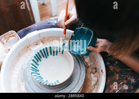 Professionelle Töpfer arbeitet auf Malplatten in der Werkstatt. Frau malt eine Keramikplatte mit einem Pinsel und blauer Farbe Stockfoto