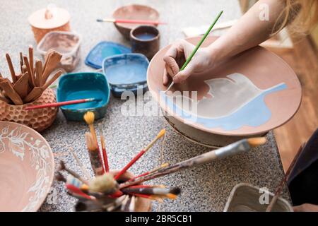 Potter Frau malt eine Keramikplatte. Mädchen zeichnet mit einem Pinsel auf Steingut. Prozess der Herstellung von Tonprodukten Stockfoto