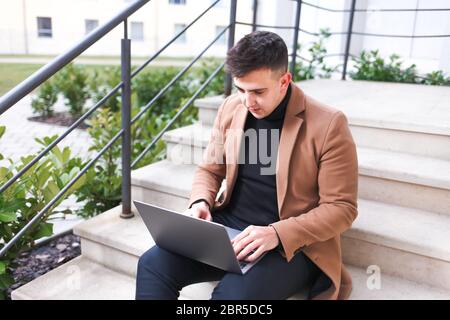 Mann in einem braunen Mantel mit Laptop in den Händen sitzt auf der Treppe. Der Student arbeitet am Computer Stockfoto