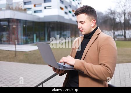 Ein Mann im Mantel hält einen Laptop in den Händen, während er vor dem Hintergrund eines Universitätsgebäudes steht. Student arbeitet auf Laptop in der Straße Stockfoto