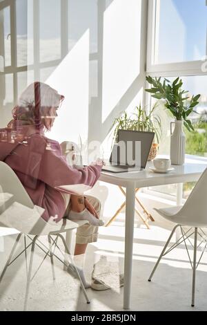 Teen Mädchen rosa Haare mit Laptop-Computer sitzen am Küchentisch hinter Glas. Stockfoto