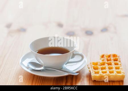 Weiße Tasse mit Untertasse und Löffel sowie angrenzende gebissene Waffel auf Holztisch in niedriger Winkel Ansicht. Stockfoto