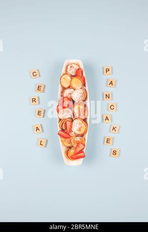 Vertikale Zusammensetzung mit Haferflocken Mini Pfannkuchen Müsli mit Erdbeeren auf weißem Teller zum Frühstück und Holzbuchstaben auf blauem Hintergrund, neuer Trend Stockfoto