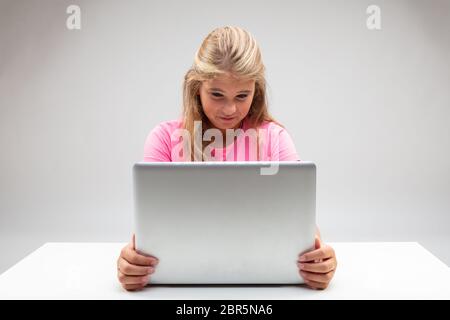 Junge blonde Mädchen, die ihren Laptop mit abfallender Grimacing in Abscheu betrachtet, als sie den Bildschirm mit beiden Händen an einem kleinen weißen Tisch sitzt hält Stockfoto