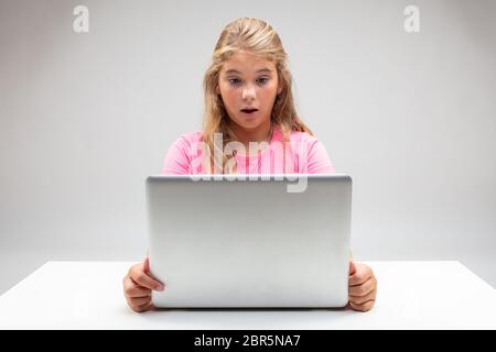 Kleine blonde Mädchen starrt auf ihren Laptop in Erstaunen mit ihren Augen weit und Mund offen überrascht, als sie an einem weißen Tisch über einem Studio backg sitzt Stockfoto