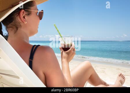 Junge Frau mit Hut sitzen auf weißen Liegestuhl genießen den Saft trinken am Strand. Stockfoto