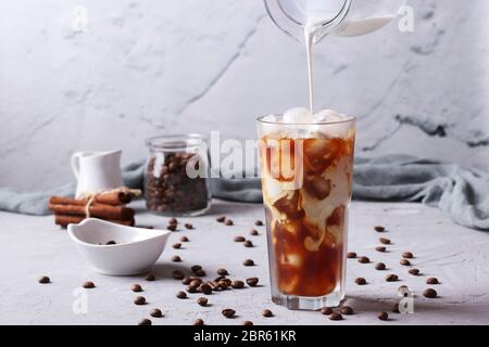 Kalter Kaffee in einem hohen Glas mit Sahne, die in ihn gegossen wird, zeigt die Textur des Getränks. Hellgrauer Hintergrund, Horizontales Format, Nahaufnahme Stockfoto