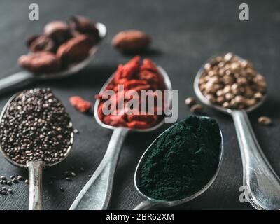 Löffel verschiedener Superfoods auf schwarzen Holztisch Hintergrund. Superfood Nahaufnahme, wie Chia Samen, Spirulina, Kakaobohne, Gojibeeren, Hanf samen Stockfoto