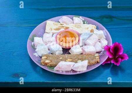 Griechischer süßer Stab von Lefkada, kandierte Früchte, Halva, türkischer Genuss in Glasplatte auf türkisfarbenem Tisch mit rosa Blume serviert Stockfoto