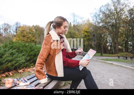 Mädchen Künstler malt ein Bild auf Leinwand direkt im Park auf einer Bank. Frau zeichnet ein Bild, das sie in den Händen hält Stockfoto