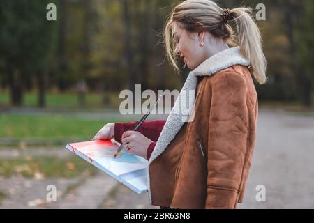 Das junge Mädchen zeichnet ein Bild direkt im Park. Malerin in einer braunen Jacke steht in einem Park und hält ein Gemälde in Händen und zeichnet sie Stockfoto