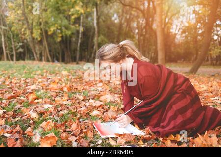 Junge Künstlerin zeichnet ein Bild kniet in einem Park auf dem Gras. Girl nimmt Inspiration für Kreativität sitzen im Herbst Park und malen Stockfoto