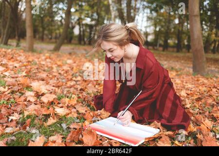 Junge Künstlerin zeichnet ein Bild kniet in einem Park auf dem Gras. Girl nimmt Inspiration für Kreativität sitzen im Herbst Park und malen Stockfoto