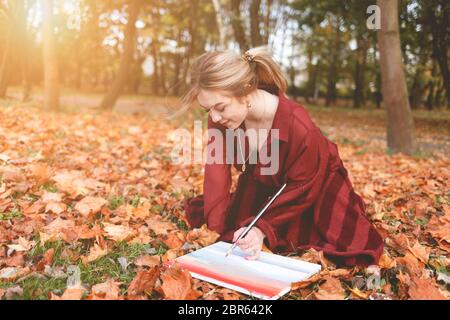 Junge Künstlerin zeichnet ein Bild kniet in einem Park auf dem Gras. Girl nimmt Inspiration für Kreativität sitzen im Herbst Park und malen Stockfoto