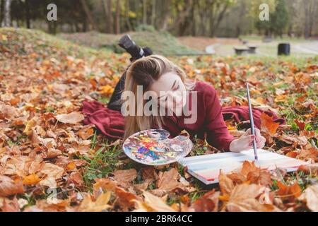 Der junge Künstler liegt im Herbstpark auf einem Plaid und malt ein Bild. Mädchen zeichnet ein Bild im Park Stockfoto