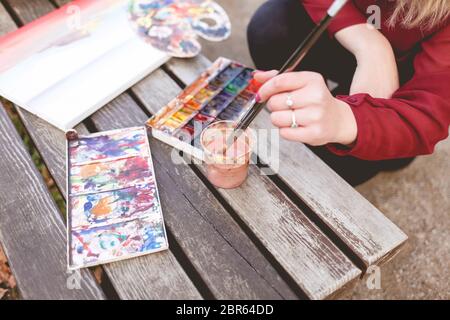 Frau zeichnet ein Bild auf einer Parkbank. Blonde Künstlerin engagiert sich in ihrem Lieblings-Hobby des Zeichnens. Zeichenzubehör Stockfoto